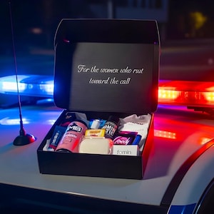 Op de afbeelding: Een zwarte geschenkdoos gevuld met diverse items, rustend op de motorkap van een politieauto met zwaailichten. Op het deksel van de doos staat: "Voor de vrouwen die naar de oproep rennen." De doos bevat een verscheidenheid aan producten, waaronder een roze spuitfles en een stuk zeep.