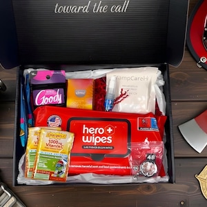 Op de afbeelding: Een cadeaupakket met items met een brandweerthema. Inclusief een rode brandweerhelm, een bijl, een Amerikaanse vlag en een doos gevuld met diverse benodigdheden zoals doekjes, vitamines en andere benodigdheden. De tekst "toward the call" is zichtbaar.