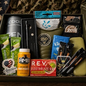 May include: A gift box containing a variety of items such as a cooling towel, energy drinks, body powder, electrolyte gum, and other essentials. The box also includes a torch, hair ties, tissues, and coffee sachets. An American flag is visible in the background.