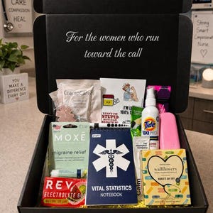 Puede incluir: Una caja de regalo negra llena de varios artículos, incluyendo un cuaderno, detergente para ropa Tide y alivio para la migraña. La caja tiene el texto "For the women who run toward the call".