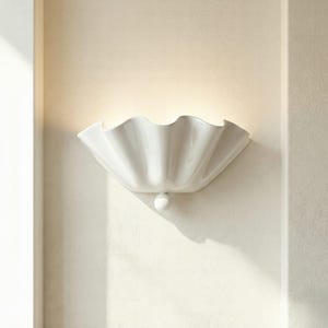 May include: A white ceramic wall sconce with a shell-shaped design. The light fixture is mounted on a cream-coloured wall, casting a soft glow. The sconce has a small, round decorative element below.