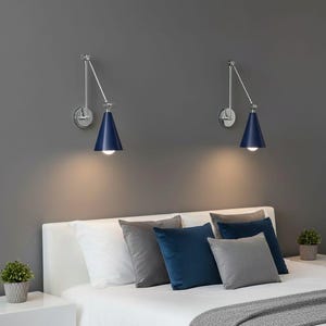 Puede incluir: Dos lámparas de pared con pantallas cónicas azul marino y brazos cromados ajustables están colocadas sobre una cama. Las lámparas están montadas en una pared gris. La cama tiene sábanas blancas y almohadas en tonos grises y azules.