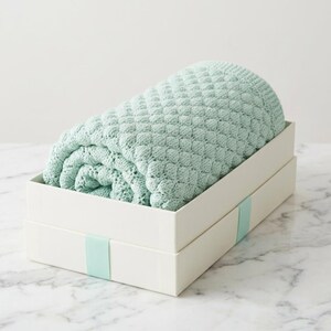 Könnte beinhalten: Eine gerollte, hellgrüne Strickdecke liegt in einer weißen rechteckigen Box. Die Decke hat ein strukturiertes, rautenförmiges Muster. Ein hellgrünes Band umschließt die Box. Die Box steht auf einer weißen Marmoroberfläche.