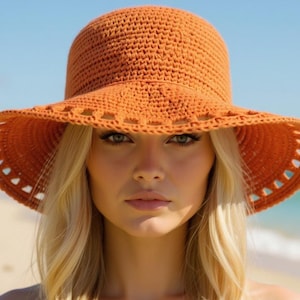 Op de afbeelding: Een close-up van een vrouw met een gehaakte oranje zonnehoed met een brede rand. De hoed heeft een ronde kroon en een decoratief opengewerkt patroon langs de rand van de rand. De achtergrond is een strandscène met blauwe lucht en oceaan.