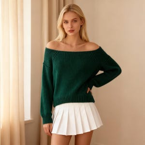 May include: A woman wearing a dark green off-the-shoulder sweater and a white pleated skirt. The sweater has long sleeves and a ribbed texture. The skirt is short and has a classic pleated design. The outfit is simple and stylish.
