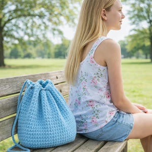 Puede incluir: Bolso de ganchillo azul claro con cordón y patrón texturizado. El bolso tiene forma redondeada y cierre de cordón. El bolso está sobre un banco de madera. Dimensiones: aproximadamente 30 cm de altura.