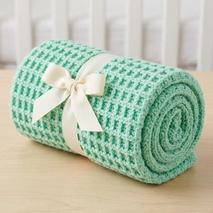 May include: Rolled-up, mint green waffle-knit blanket tied with a cream-colored ribbon and bow. The blanket is made of a soft, textured material and is presented on a light wooden surface. The background includes a white crib.
