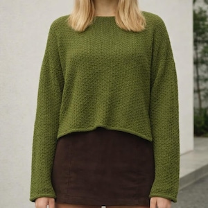Peut inclure: Un pull vert à manches longues avec un motif en tricot texturé. Le pull a un col rond et une coupe courte. Le modèle porte une jupe marron. Le pull est de couleur unie et semble être fait d'une matière douce.