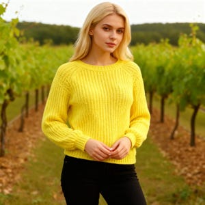 May include: A bright yellow sweater with a ribbed knit texture. The sweater has a crew neck and long sleeves. The person in the image is wearing black pants. The background is a vineyard with rows of grapevines.