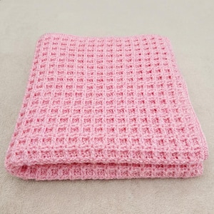 Peut inclure: Une couverture tricotée rose clair pliée. La couverture présente une texture gaufrée, avec un motif répétitif de petits carrés. Les bords sont soigneusement finis, et la couverture semble douce et confortable.