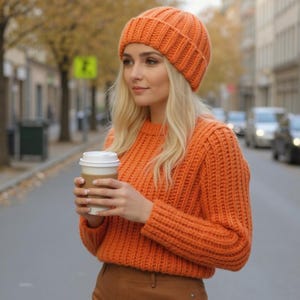May include: An orange knit beanie and sweater set. The beanie is a ribbed knit design. The sweater has a crew neck and long sleeves. The person is holding a coffee cup with a white lid. The background is a city street with cars and trees.