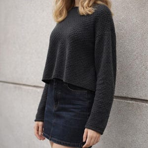Könnte beinhalten: Ein dunkelgrauer, strukturierter, gecroppter Pullover mit langen Ärmeln. Dazu wird ein dunkler Jeans-Minirock getragen. Das Outfit ist lässig und alltagstauglich. Die Textur des Pullovers sorgt für optisches Interesse.