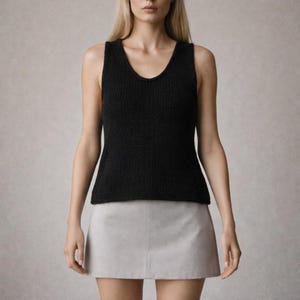 May include: A black tank top with a V-neck design. The top is made of a textured knit fabric. The model is wearing a light gray mini skirt. The background is a solid light gray.