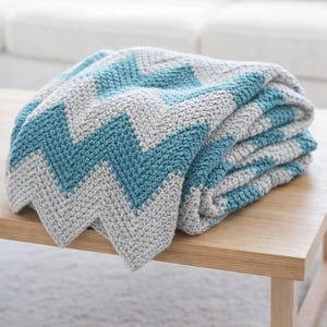 May include: A folded crochet blanket with a chevron pattern in grey and teal blue. The blanket, made from thick yarn, is neatly folded on a wooden table. The design features a repeating zigzag pattern, offering a cosy aesthetic.