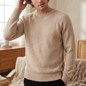 May include: A beige crew neck jumper with raglan sleeves. The jumper has a ribbed hem and cuffs. The material appears to be a soft knit. The jumper is a neutral colour and suitable for various outfits.