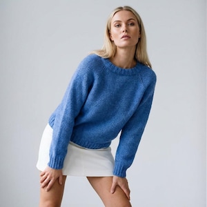 Pode incluir: Um suéter azul de gola redonda com mangas compridas. O suéter é combinado com uma saia branca. A modelo está inclinada para a frente com as mãos nas pernas. O fundo é uma parede branca lisa.