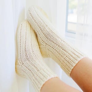 May include: A pair of cream-colored knitted socks with ribbed cuffs and decorative vertical patterns. The socks are resting against a white curtain, suggesting a cozy and comfortable setting. The socks appear to be handmade.