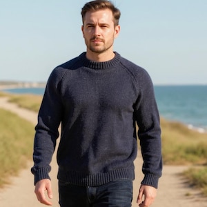 May include: A navy blue knitted sweater with a crew neck and ribbed cuffs and hem. The person in the image is walking on a path near the sea. The sweater is made of a textured knit fabric.