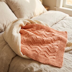 May include: A coral-colored knitted blanket with a chevron pattern is draped over a bed. The blanket has a textured design and a solid border. A cream-colored, lightweight blanket is layered underneath.