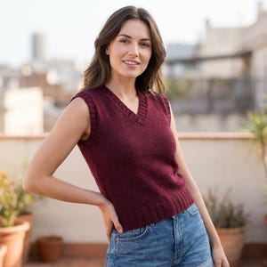 Könnte beinhalten: Eine weinrote ärmellose V-Ausschnitt-Weste. Das Strickware hat einen gerippten Saum und eine klassische Passform. Das Model trägt blaue Jeansshorts. Der Hintergrund zeigt eine Dachterrasse mit Topfpflanzen und einer verschwommenen Stadtlandschaft.