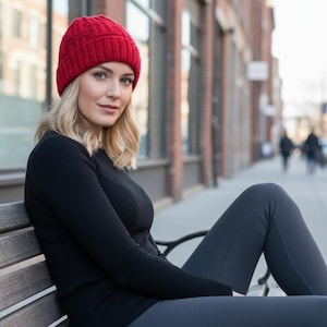 May include: A woman wearing a red knit beanie, black long-sleeve shirt, and gray leggings sits on a wooden bench. The background shows a brick building and a city street.