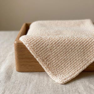 May include: A cream-colored, knitted square cloth is neatly folded and placed inside a wooden box. The cloth has a textured appearance, with visible knit stitches. The box is made of light-colored wood, and the background is a neutral tone.