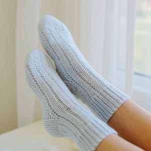 Può includere: Un paio di calzini lavorati a maglia azzurri con un design a coste e dettagli traforati. I calzini sono indossati sui piedi di una persona, con le gambe distese. I calzini sembrano morbidi e confortevoli.