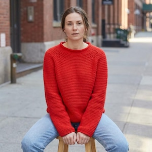 May include: A bright red knit sweater with a crew neck. The sweater is paired with light blue jeans. The person is sitting on a wooden stool outdoors, with a brick building in the background.