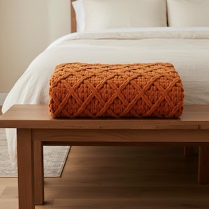 Op de afbeelding: Een oranje, dikgebreide sprei met een ruitpatroon ligt op een houten bank. De sprei is rechthoekig en ziet er zacht uit. De achtergrond toont een bed met wit beddengoed en een neutrale muur.