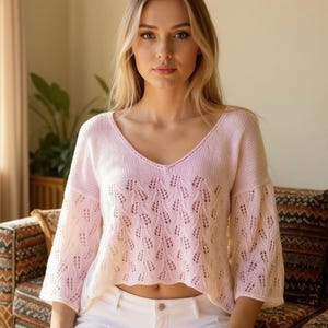 Peut inclure: Un pull rose clair à mailles ajourées avec un col en V et des manches trois-quarts. Le pull présente un motif en forme de feuille et est porté avec un pantalon blanc. Le modèle est assis sur un canapé à motifs.