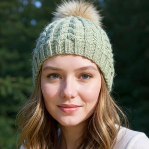 Può includere: Un berretto lavorato a maglia verde chiaro con un motivo a trecce e un pompon in finta pelliccia. Il cappello è realizzato in un materiale morbido e strutturato. Un accessorio invernale.