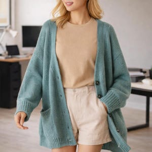 Może przedstawiać: Jasnoniebieski kardigan z dużymi guzikami i dwiema przednimi kieszeniami. Kardigan jest noszony na beżowym t-shircie i beżowych szortach. Strój jest swobodny i wygodny.