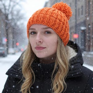 Könnte beinhalten: Eine orangefarbene Strickmütze mit Bommel. Die Person trägt eine schwarze Jacke und befindet sich in einer verschneiten Umgebung im Freien. Die Mütze hat eine warme, leuchtende Farbe.