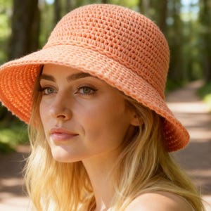 Puede incluir: Un sombrero de sol de ganchillo color melocotón con ala ancha. El sombrero está hecho de un hilo texturizado y tiene una corona redondeada. El sombrero se usa al aire libre, con un fondo borroso de árboles y un sendero.