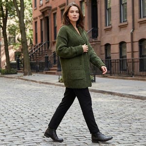 Könnte beinhalten: Eine Frau trägt einen langen, olivgrünen Cardigan mit zwei Vordertaschen, kombiniert mit einer schwarzen Hose und schwarzen Stiefeln. Der Cardigan ist grob gestrickt. Das Outfit wird beim Gehen auf einer Kopfsteinpflasterstraße getragen.