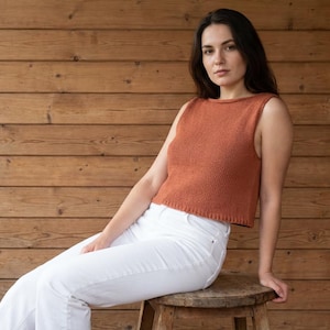 May include: A sleeveless, terracotta-coloured knitted top and white trousers. The top has a simple, round neckline and a ribbed texture. The model is seated on a wooden stool against a backdrop of wooden planks.