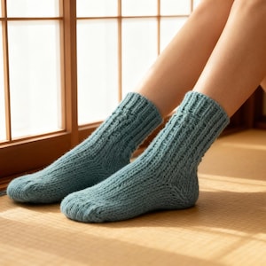 May include: A pair of teal-colored, hand-knitted socks. The socks are thick, with a ribbed pattern on the cuff and a textured knit throughout the body. The socks are displayed indoors, near a window, suggesting a cozy, comfortable theme.