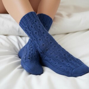 May include: A pair of blue knitted socks with a textured cable pattern. The socks are folded over each other, resting on a white bed sheet. The socks are a solid, deep blue colour.