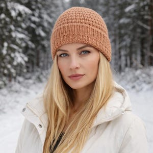 May include: A woman wearing a tan knitted beanie hat and a cream-colored jacket. The hat has a ribbed brim and a textured crown. The background shows a snowy forest, suggesting a winter setting. The woman has long blonde hair and blue eyes.