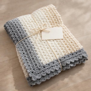 Puede incluir: Una manta de ganchillo hecha a mano en crema, blanco y gris, atada con hilo y una etiqueta en blanco. La manta presenta un patrón de puntadas texturizadas y un borde gris festoneado. Una manta acogedora, suave y cálida.
