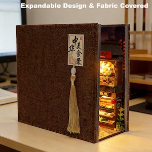 Könnte beinhalten: Ein stoffbezogenes, erweiterbares, buchförmiges Miniatur-Diorama. Das offene Buch zeigt eine detaillierte, beleuchtete Miniatur-Szene eines asiatischen Restaurants. Der Einband hat ein dekoratives Etikett mit chinesischen Schriftzeichen und eine Quaste.