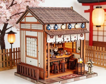 May include: A miniature Japanese-style bar with a brown wooden exterior, featuring a detailed interior with a bar, stools, and bottles. The bar has a brown tiled roof, decorative lanterns, and a small figure behind the bar. Cherry blossoms and a traditional gate are in the background.