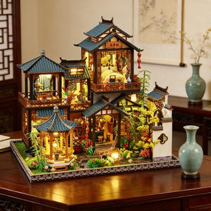 Puede incluir: Una detallada casa en miniatura de estilo asiático con múltiples niveles, con tejados de tejas azules, intrincadas estructuras de madera e iluminación interior. El modelo incluye pequeñas figuras, linternas decorativas y vegetación circundante.