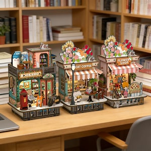 May include: Three miniature wooden shop models: a bookstore, a cat coffee shop, and a flower shop. The bookstore has a red mailbox and a bicycle. The cat coffee shop has a street lamp. The flower shop has a striped awning. Each model has a detailed design.