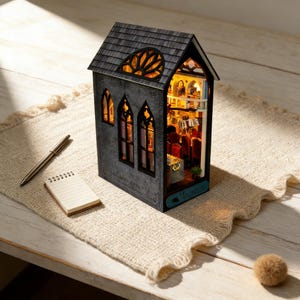 May include: A miniature, illuminated gothic-style house with a gray exterior and a detailed interior. The house features arched windows, a decorative roof, and a small sign with text. A pen and notepad are next to the house.