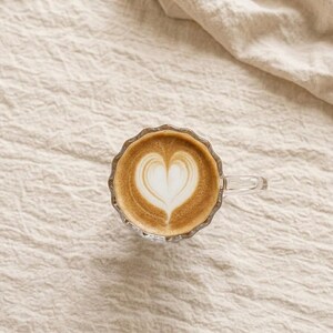 May include: A glass cup of coffee with a heart-shaped design in the foam. The coffee is light brown, and the foam is white. The cup has a scalloped edge and a handle. The cup is on a beige, textured surface.