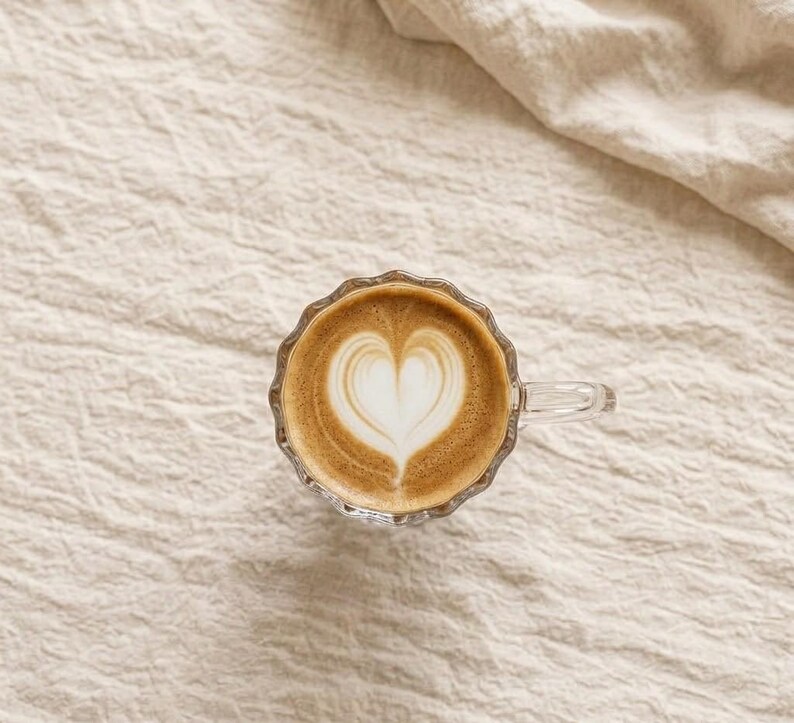 May include: A glass cup of coffee with a heart-shaped design in the foam. The coffee is light brown, and the foam is white. The cup has a scalloped edge and a handle. The cup is on a beige, textured surface.