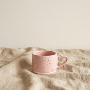May include: A pink ceramic mug with a heart-shaped handle. The mug has a speckled design with small black dots and a white base. The mug is sitting on a beige linen surface.