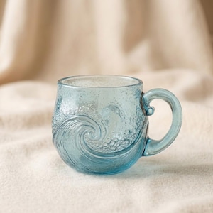 May include: A light blue glass mug with a wave design. The mug has a rounded body and a curved handle. The wave design is embossed on the side of the mug. The mug is sitting on a white surface.