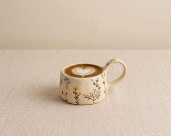 Puede incluir: Una taza de cerámica beige con un diseño de arte latte en forma de corazón. La taza está decorada con patrones florales coloridos y tiene un asa curva. La taza está llena de café y se encuentra sobre una superficie de color neutro.